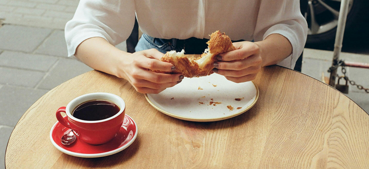 Woman enjoying a croissant and coffee at a café, representing a moment of relaxation and wellness.