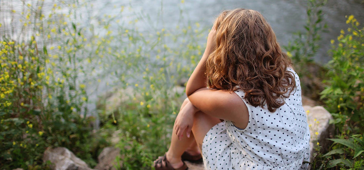 Woman sat by a lake, holding her head in her hands, because of the impact of hormonal stress.