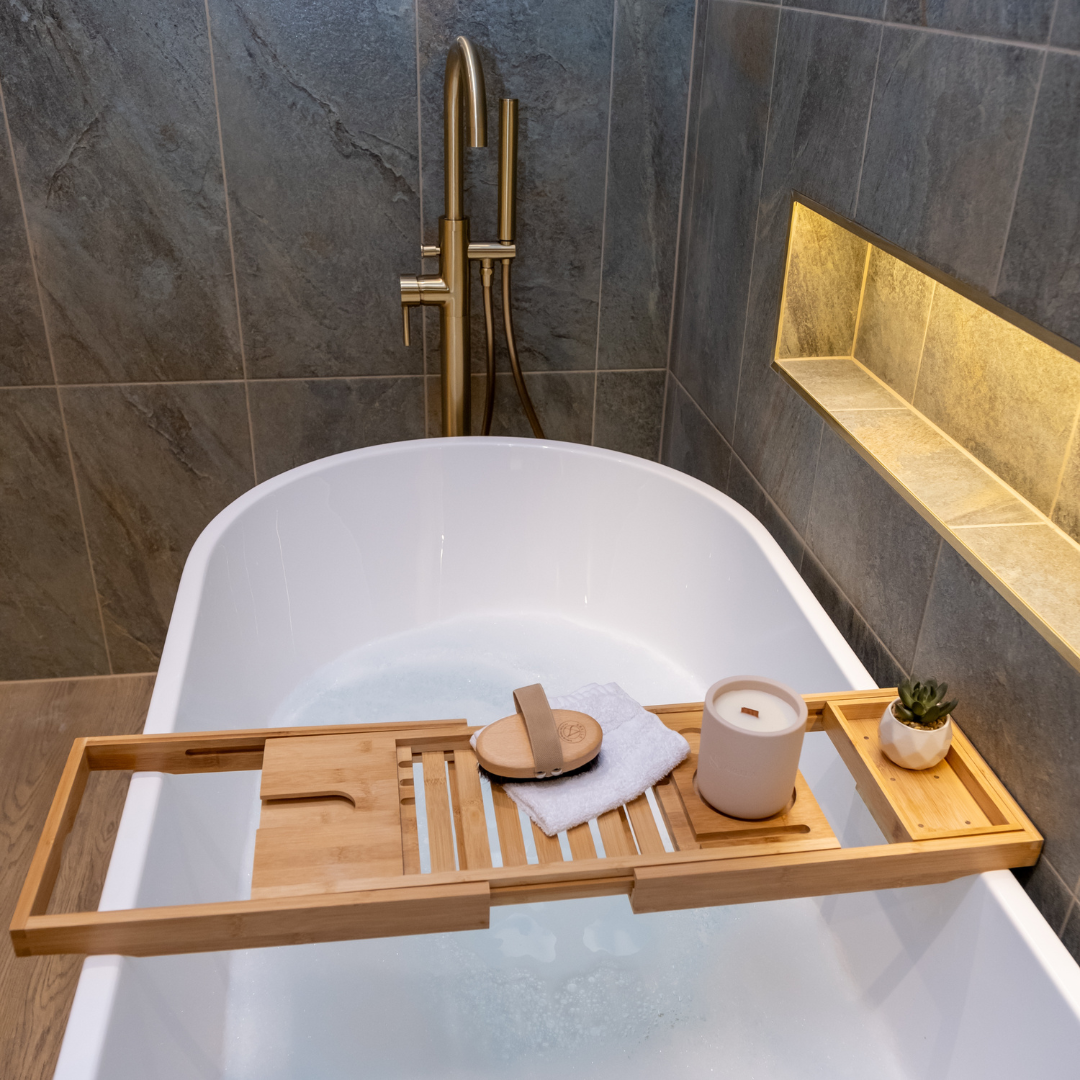 Elegant bathroom with a bathtub, gold taps and wooden tray holding a candle, body brush and plant.